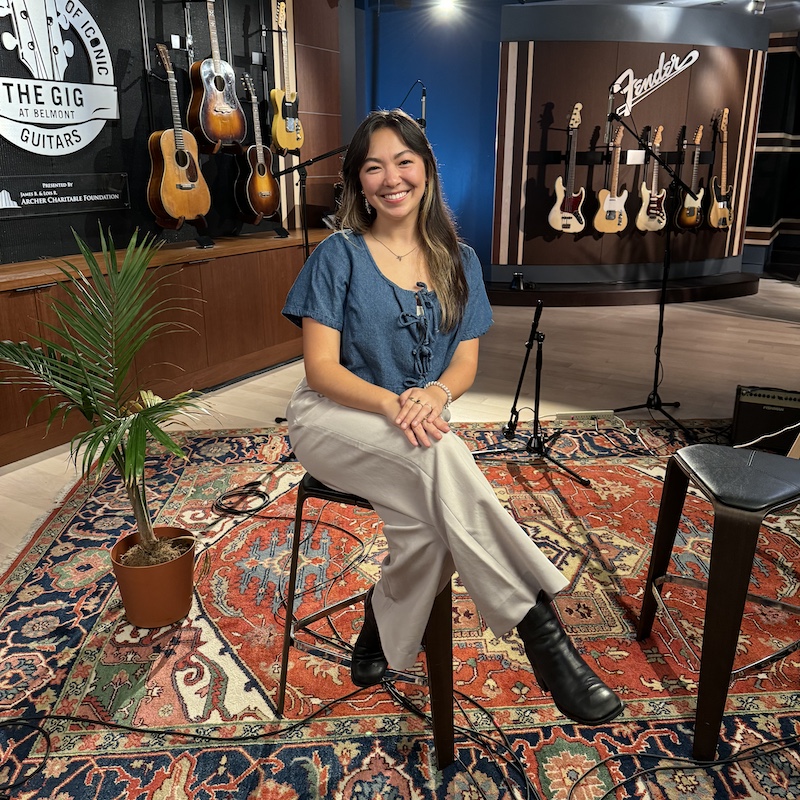 female student Calista Morrison sitting on a stool in a carpeted room with guitars in the background