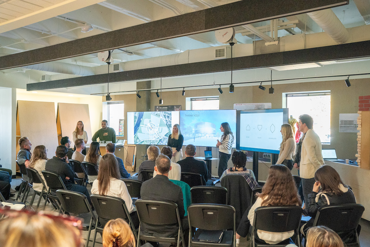Students present architectural designs to a seated audience using digital renderings and large display monitors.