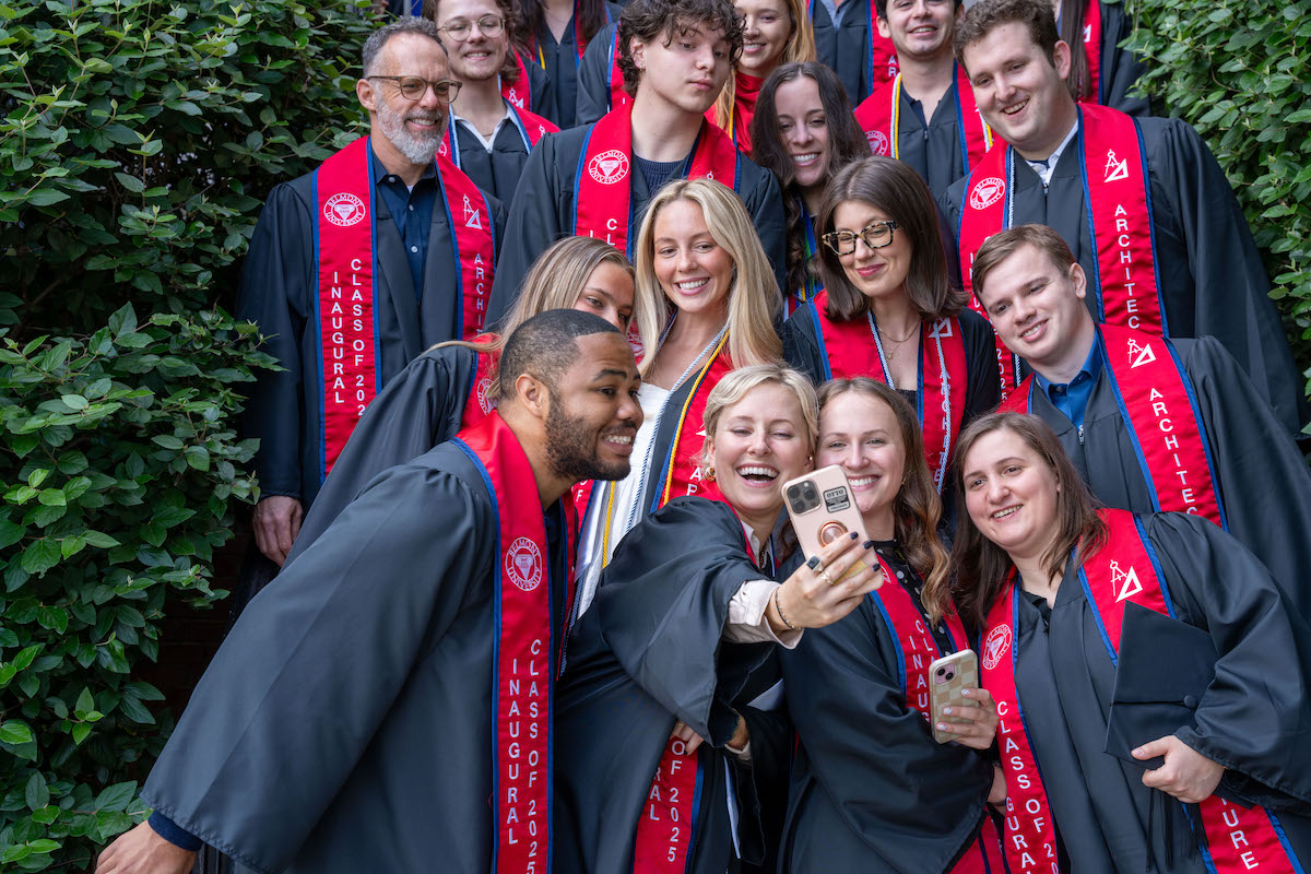 Graduates in regalia and “Class of 2025” stoles smile and pose for a celebratory group selfie outdoors.