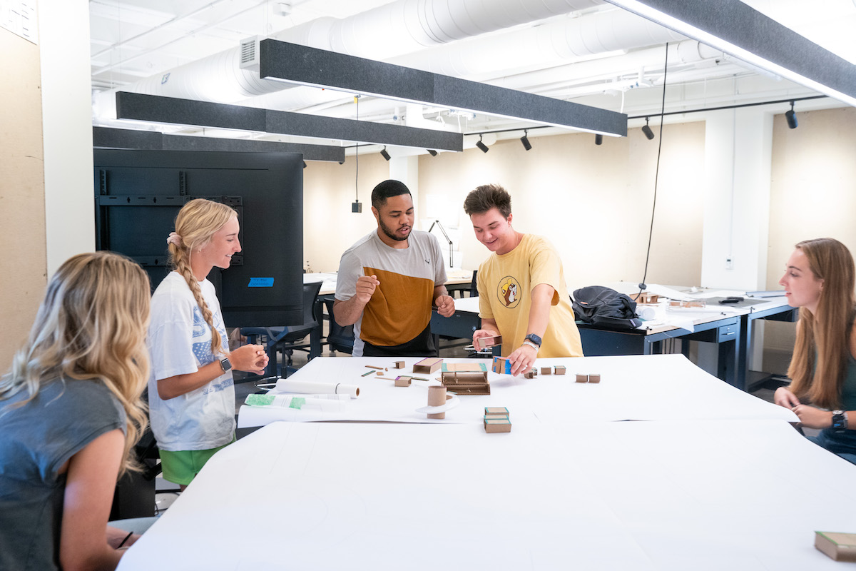 Architecture students collaborate around a table, building models and discussing ideas during a hands-on design studio session.
