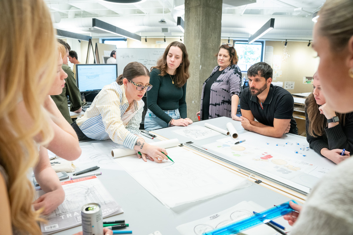 Students and faculty gather around a worktable as one student sketches ideas on architectural layout paper.