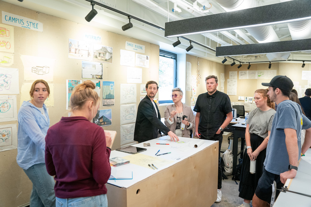 Architecture students engage in critique and discussion with visiting reviewers in a classroom filled with sketches and diagrams.