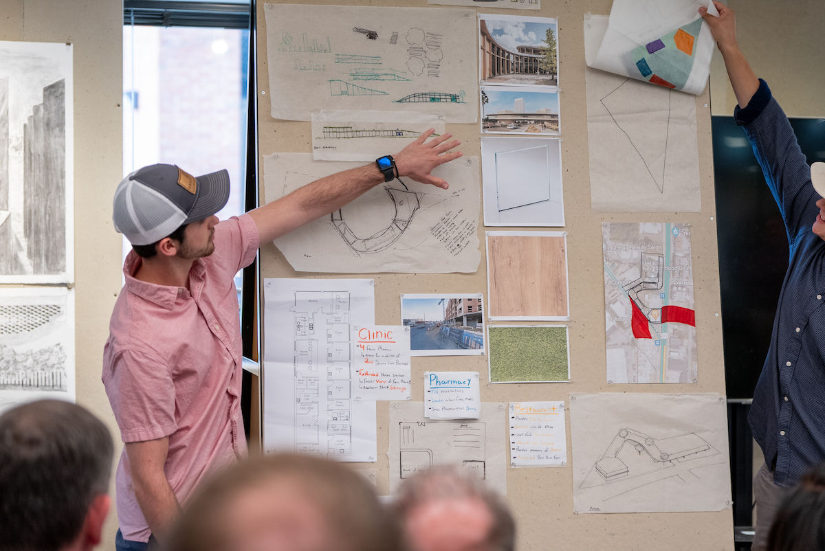 Student gestures toward concept drawings and materials pinned to a wall during an architecture critique session.