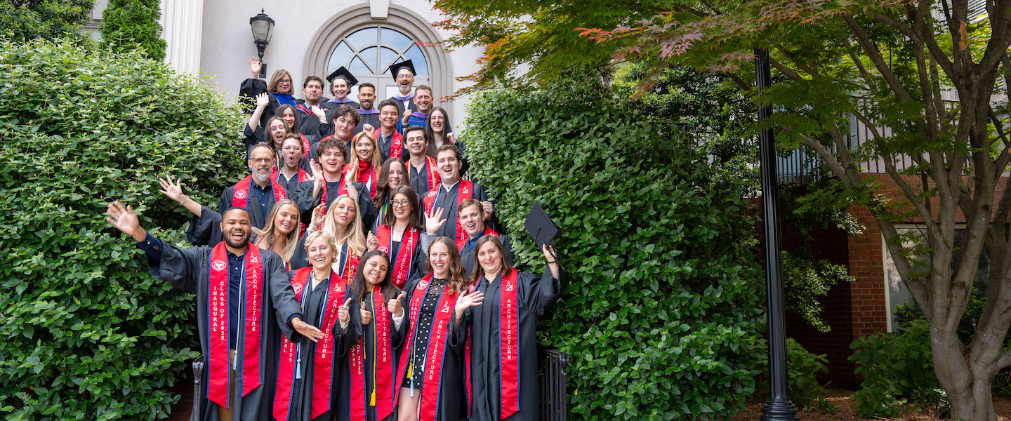 Graduating architecture students and faculty pose together in front of a campus building, celebrating in caps, gowns, and red stoles.