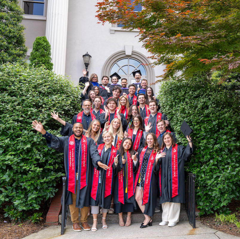 Graduating architecture students and faculty pose together in front of a campus building, celebrating in caps, gowns, and red stoles.