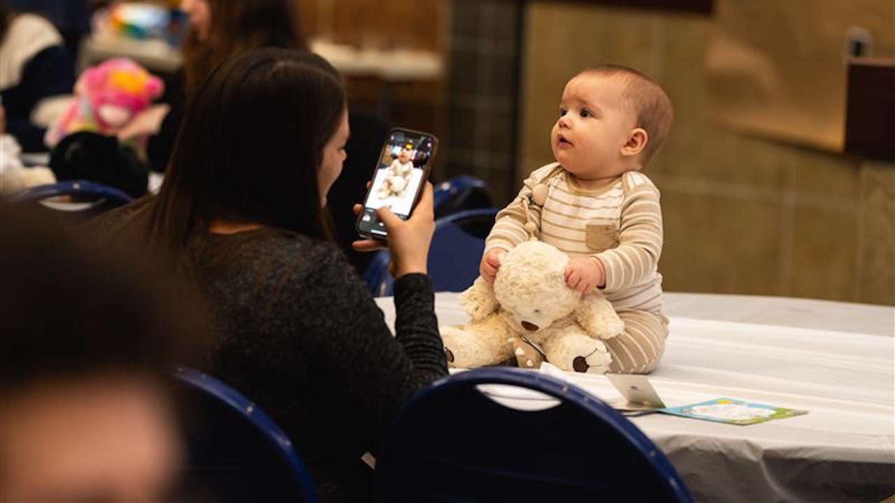 mother taking a photo on her phone of a baby sitting on a table with a stuffed bruin