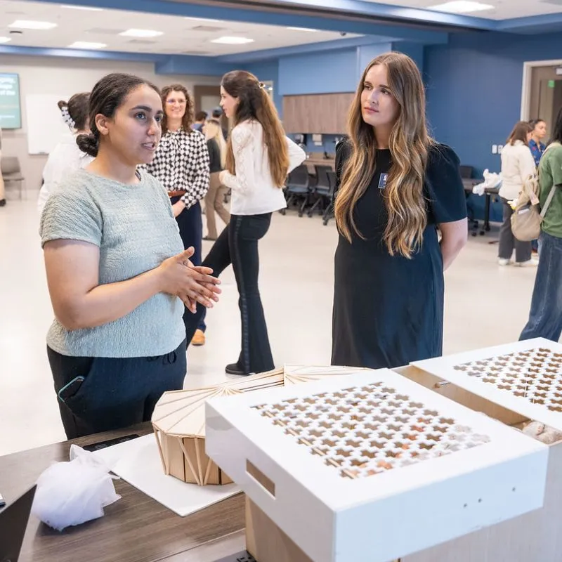 An architecture student explains her design for life-saving baby care boxes to a nursing student during a collaborative presentation at Belmont University. The setting features a modern classroom with other students engaged in discussions in the background, highlighting the partnership between the two disciplines.