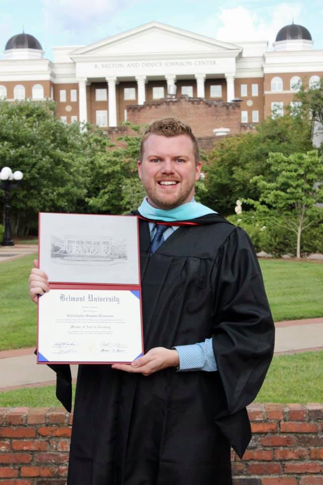 A graduate in academic regalia with teal hood displays a diploma in front of Belmont University's Johnson Center, a brick building with white columns and black domes.