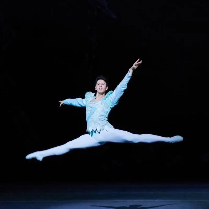 A male ballet dancer performs a grand jeté in mid-air, wearing a light blue costume and white tights, against a dark background.