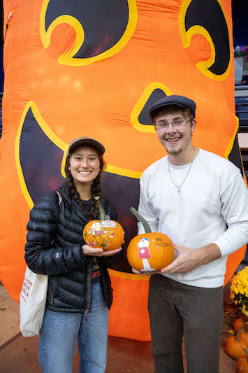 Two students holding pumpkins in front of giant pumpkin inflatable