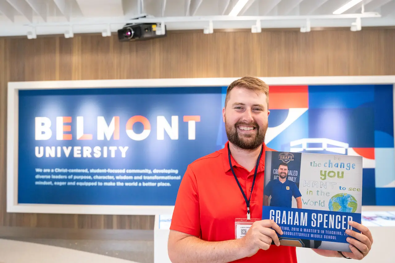 A man with a beard wearing a red shirt and name badge smiles while holding a promotional card in front of a Belmont University branded wall display.