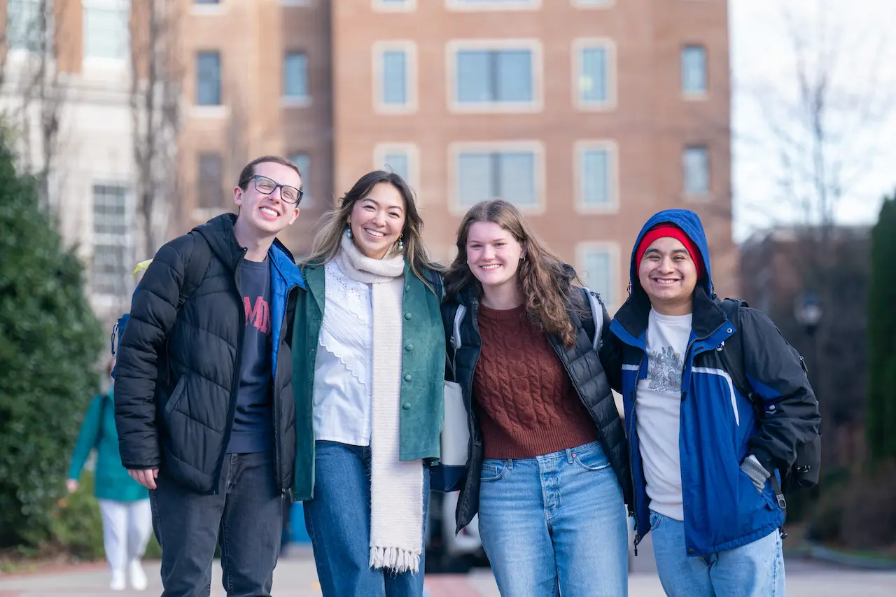 Four students smile while standing together on a college campus pathway with a brick building in the background. They are wearing winter coats and standing side by side.