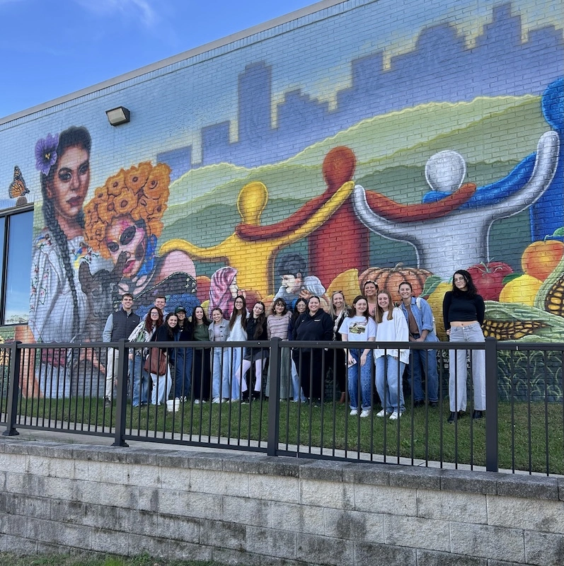 A diverse group of people posing in front of a colorful mural depicting community and cultural themes, with a bright blue sky overhead.