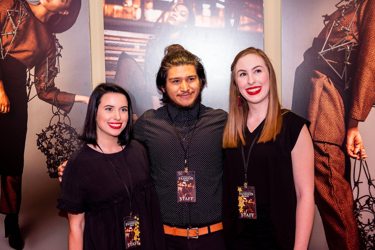 Three smiling people with staff passes in front of fashion show posters at the event.