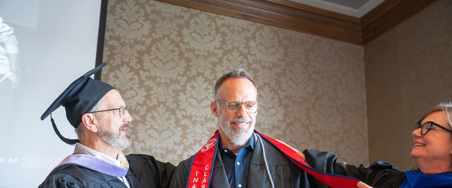 The image shows a graduation ceremony moment with three people in an indoor setting. On the left, a person wearing academic regalia including a graduation cap and gown with purple trim appears to be congratulating or presenting something to a middle-aged man with glasses and a gray beard. The man in the center is wearing a dark shirt with a red and blue academic stole or sash that has text on it. On the right, a woman with glasses is smiling at the interaction. The background features ornate beige patterned wallpaper and what appears to be a projection screen partially visible on the left edge of the image.