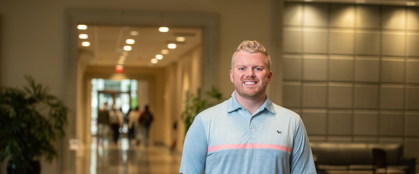 A young educator stands smiling in a university hallway. He wears a light blue polo shirt with a pink horizontal stripe and has blonde curly hair. Behind him, the hallway features modern lighting and potted plants, with students visible in the background