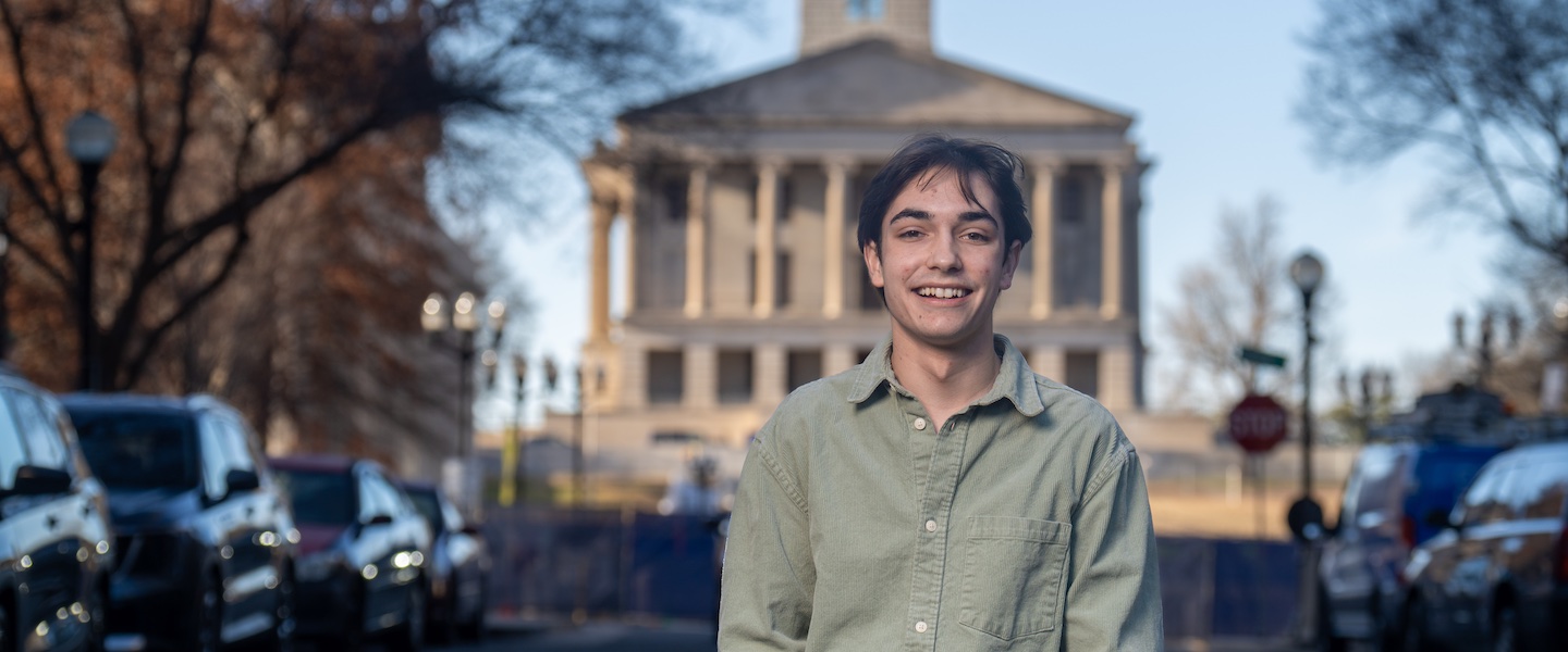 The image shows a young man smiling at the camera while standing outdoors on what appears to be a college or government building campus. He is wearing a casual light green button-down shirt. In the background is a prominent neoclassical-style building with tall columns and a symmetrical facade. The scene includes parked cars along the street and bare trees, suggesting it was taken during fall or winter. The lighting creates a soft, slightly blurred background effect while keeping the subject in sharp focus.