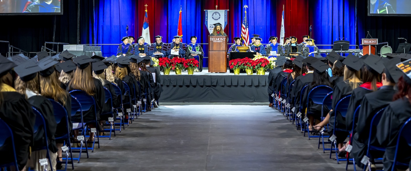 Faculty in academic regalia sit on stage behind a podium at a Belmont University ceremony as graduates face the stage.