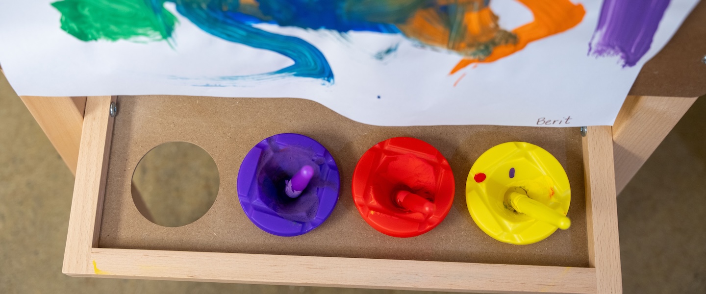 Art tray holding cups of purple, red and yellow paint with brushes beneath a colorful painting