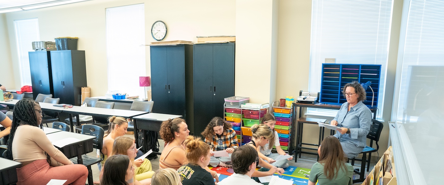 The image shows a classroom setting with a group of students sitting at desks and focused on colorful worksheets or papers. An instructor, who appears to be a woman with glasses and curly hair, stands at the front of the classroom near a whiteboard. The classroom has typical educational furniture including student desks arranged in rows, storage cabinets along the walls, colorful organizational drawers, and a wall clock. Natural light comes in through windows with blinds. The students appear engaged in their classwork while the instructor seems to be providing guidance or instruction.