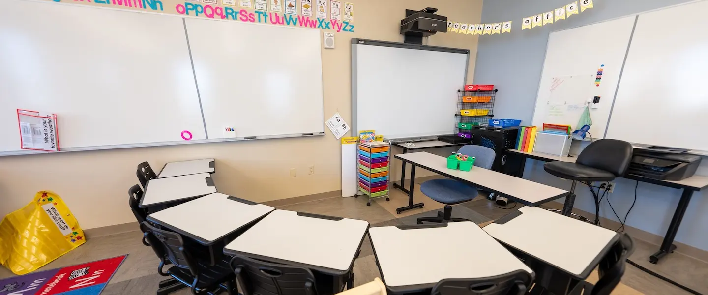 A small classroom with desks arranged in a semicircle formation. The room features whiteboards mounted on the walls, an alphabet display across the top of one wall, and a projector mounted to the ceiling. Colorful organizational elements include plastic storage bins in various colors, a rainbow-colored rolling cart with drawers, and some educational materials. The classroom has chairs for students, a teacher's desk area, and appears designed for elementary education with its bright, organized layout.
