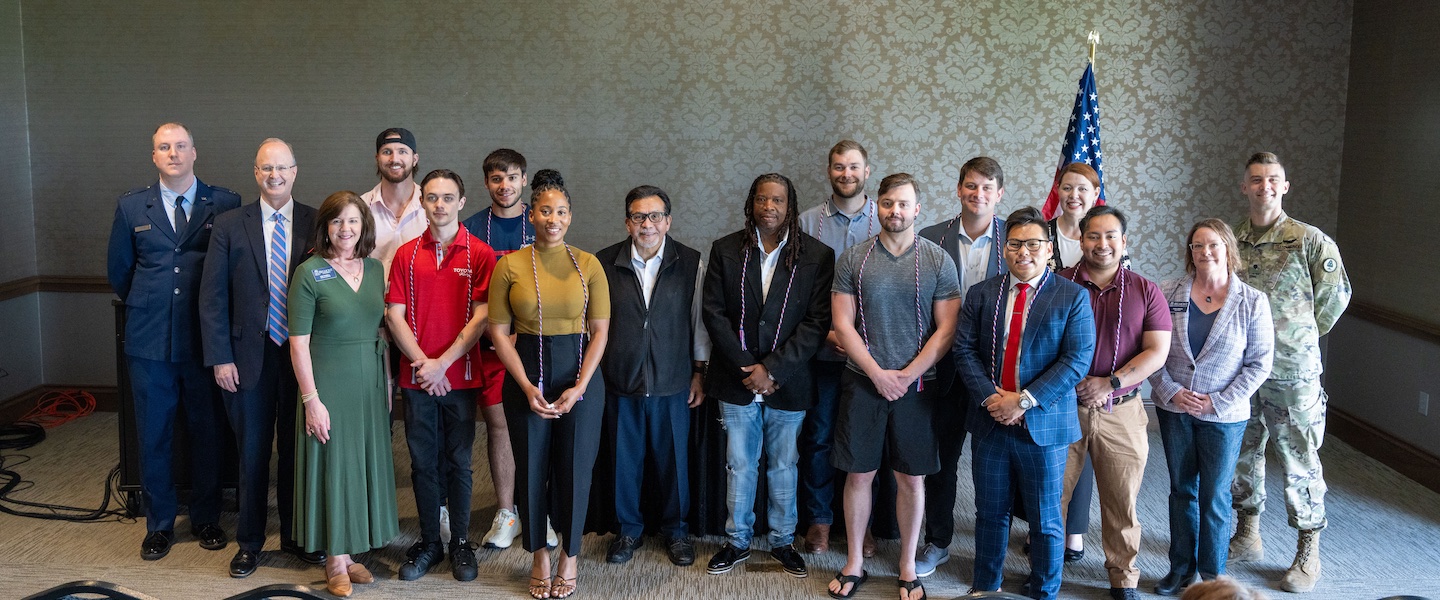 The image shows a group of graduates wearing academic honor cords standing together for a formal photograph at what appears to be a challenge coin and cord ceremony at Belmont University on Thursday, May 1.