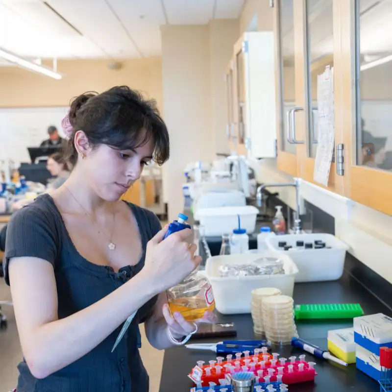 A young woman wearing a gray shirt and gloves is carefully using a pipette to transfer a liquid from a jar in a laboratory setting, with various laboratory equipment and supplies visible in the background.