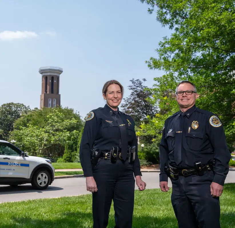 Two police officers, a woman and a man, stand smiling in a green park area with a water tower in the background. They are both wearing dark police uniforms and appear confident and approachable. A police vehicle is parked nearby.