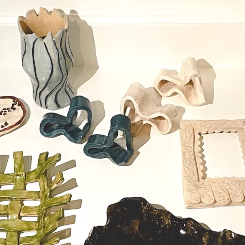 O'More students' pottery: A collection of ceramic objects displayed on a white surface, including a wavy-edged vase with black line details, abstract sculptural pieces in cream and deep blue, a textured picture frame, and a variety of uniquely shaped decorative pieces in green, black, and neutral tones.