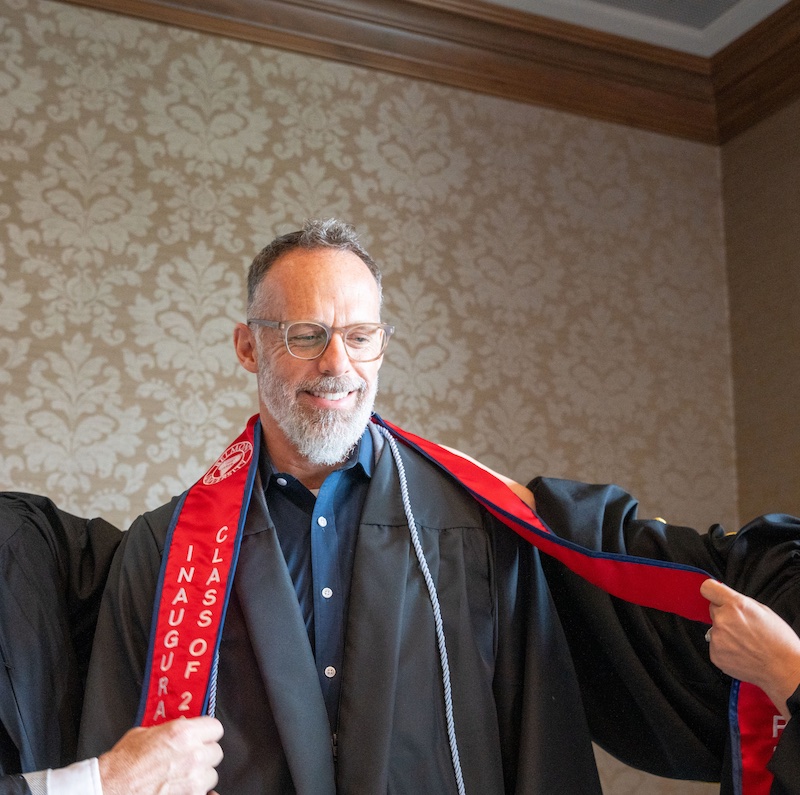 The image shows a graduation ceremony moment with three people in an indoor setting. On the left, a person wearing academic regalia including a graduation cap and gown with purple trim appears to be congratulating or presenting something to a middle-aged man with glasses and a gray beard. The man in the center is wearing a dark shirt with a red and blue academic stole or sash that has text on it. On the right, a woman with glasses is smiling at the interaction. The background features ornate beige patterned wallpaper and what appears to be a projection screen partially visible on the left edge of the image.