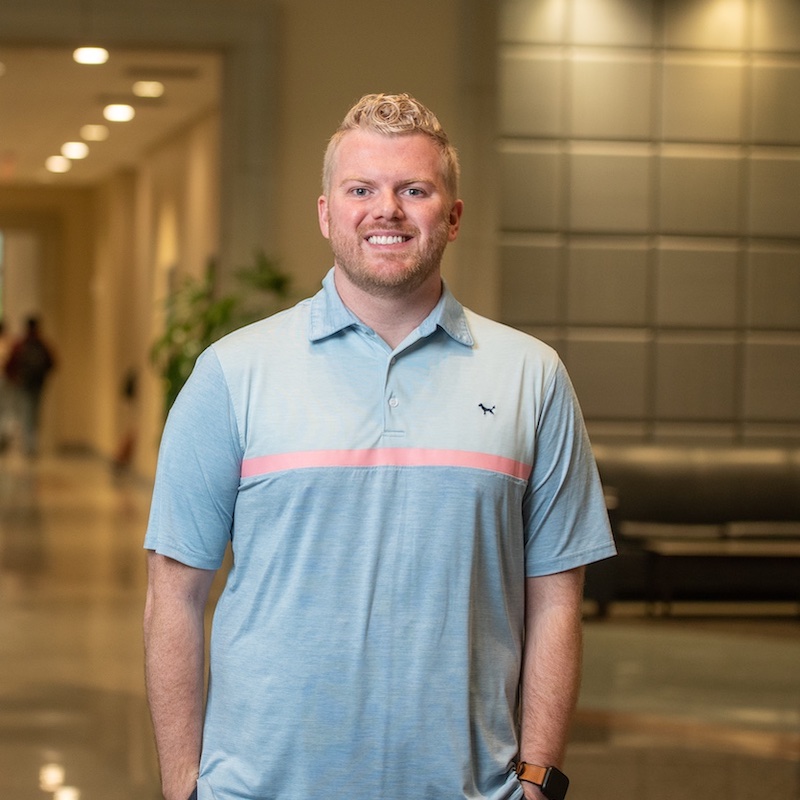 A young educator stands smiling in a university hallway. He wears a light blue polo shirt with a pink horizontal stripe and has blonde curly hair. Behind him, the hallway features modern lighting and potted plants, with students visible in the background