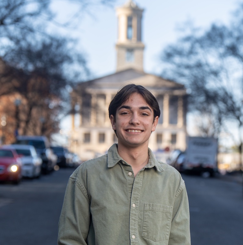 The image shows a young man smiling at the camera while standing outdoors on what appears to be a college or government building campus. He is wearing a casual light green button-down shirt. In the background is a prominent neoclassical-style building with tall columns and a symmetrical facade. The scene includes parked cars along the street and bare trees, suggesting it was taken during fall or winter. The lighting creates a soft, slightly blurred background effect while keeping the subject in sharp focus.