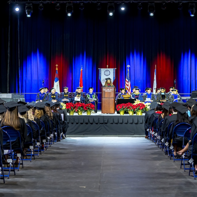 Faculty in academic regalia sit on stage behind a podium at a Belmont University ceremony as graduates face the stage.