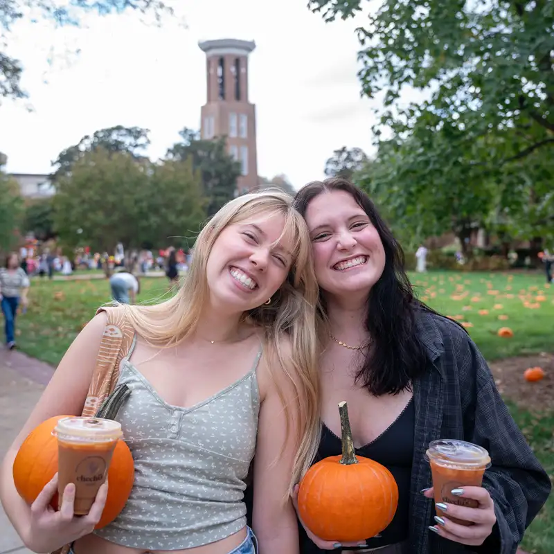 Two smiling young women pose together outdoors at Belmont University, each holding a small pumpkin. One woman has a drink in her hand, and they stand in front of the university's bell tower, with a festive atmosphere and a field of pumpkins in the background.