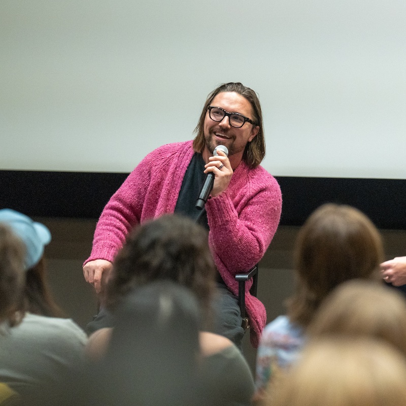 A closer shot of Gabe Simon smiling while speaking into a microphone. He has shoulder-length hair, black-framed glasses, and is wearing a vibrant pink cardigan. The audience is partially visible in the foreground, showing the backs of students' heads as they listen to his presentation.