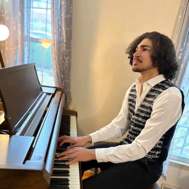 Isaac Bouldin in a patterned vest and white shirt seated at a piano, playing keys beside a window with sheer curtains