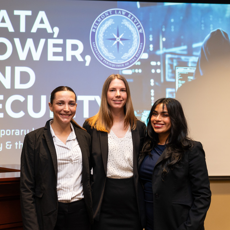 Three professionally dressed students stand together smiling in front of a screen, with the Belmont Law Review logo displayed