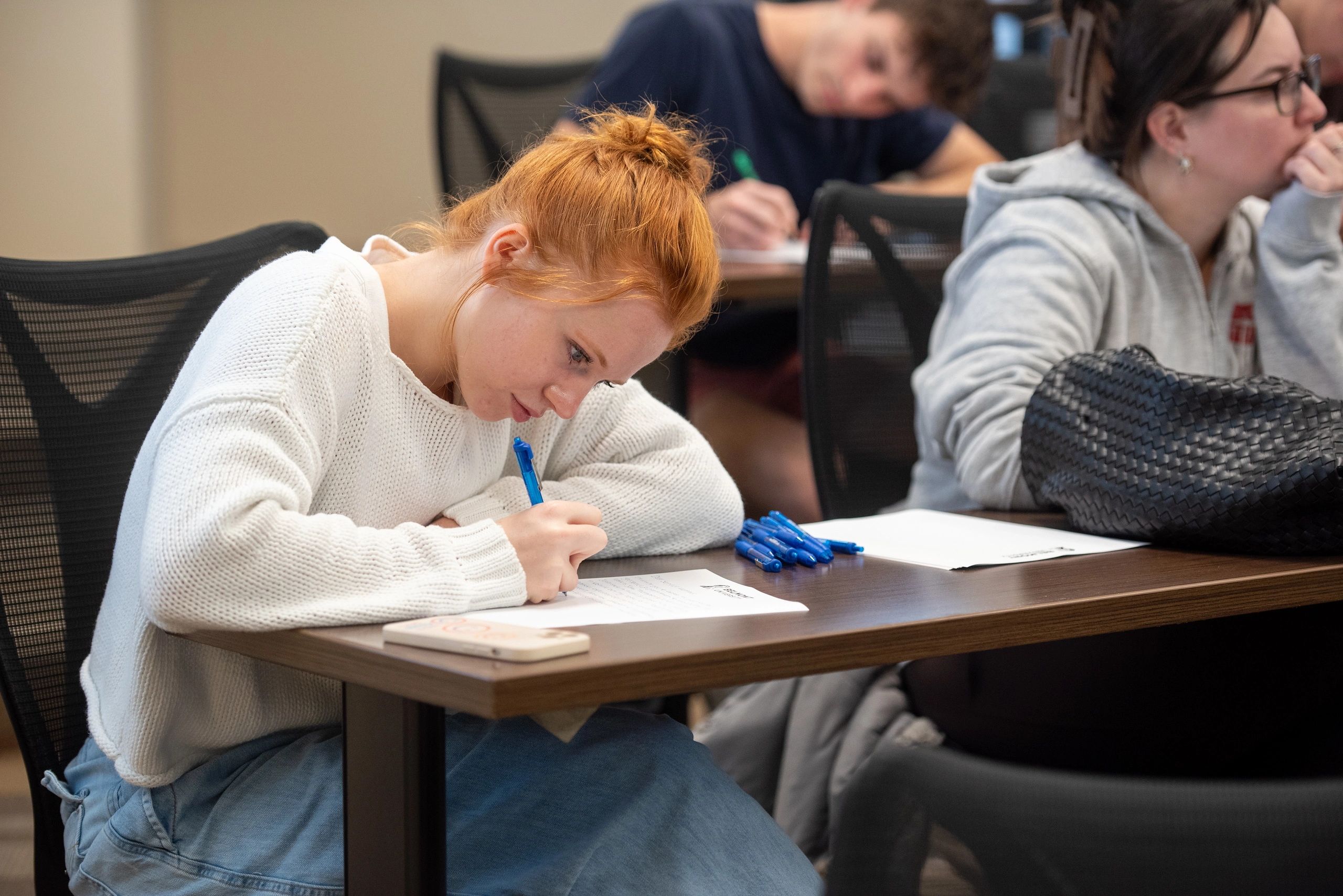 A student with red hair concentrates on writing with a blue pen during a class, leaning over a desk filled with papers and additional pens. In the background, other students are also engaged in their work, creating a focused learning environment.