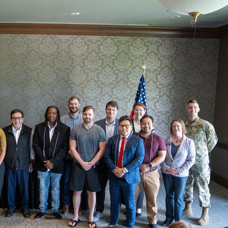 The image shows a group of graduates wearing academic honor cords standing together for a formal photograph at what appears to be a challenge coin and cord ceremony at Belmont University on Thursday, May 1.