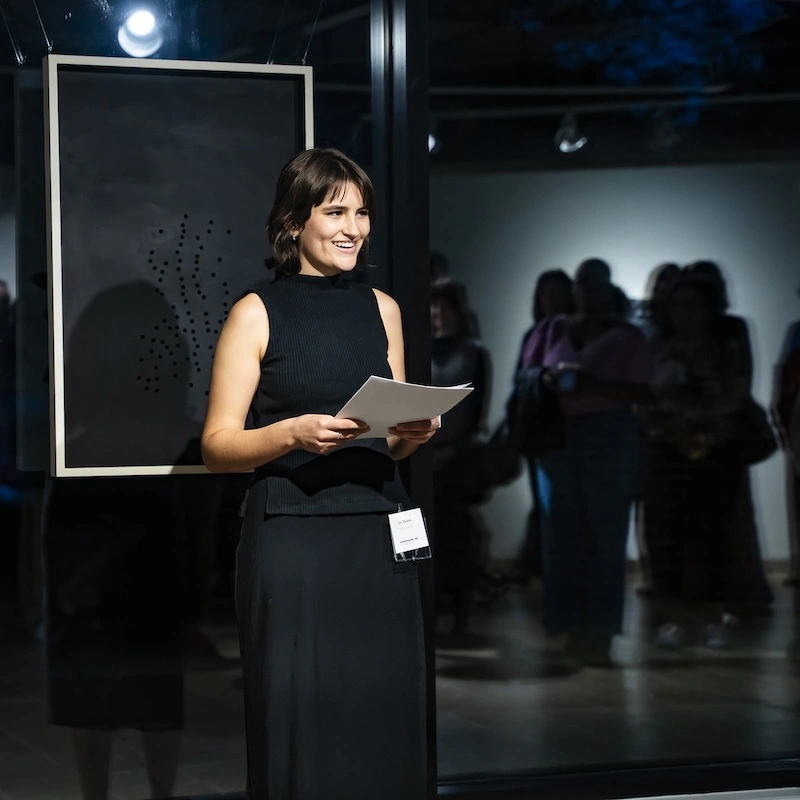 A young woman is standing in an art gallery, smiling as she holds a piece of paper in her hands. She is wearing a sleeveless black dress and appears to be presenting or speaking to an audience. Behind her, a dark artwork is displayed on the wall, and several people can be seen in the background, engaged in conversation or observing the art. The lighting is dim, creating an intimate atmosphere for the event.