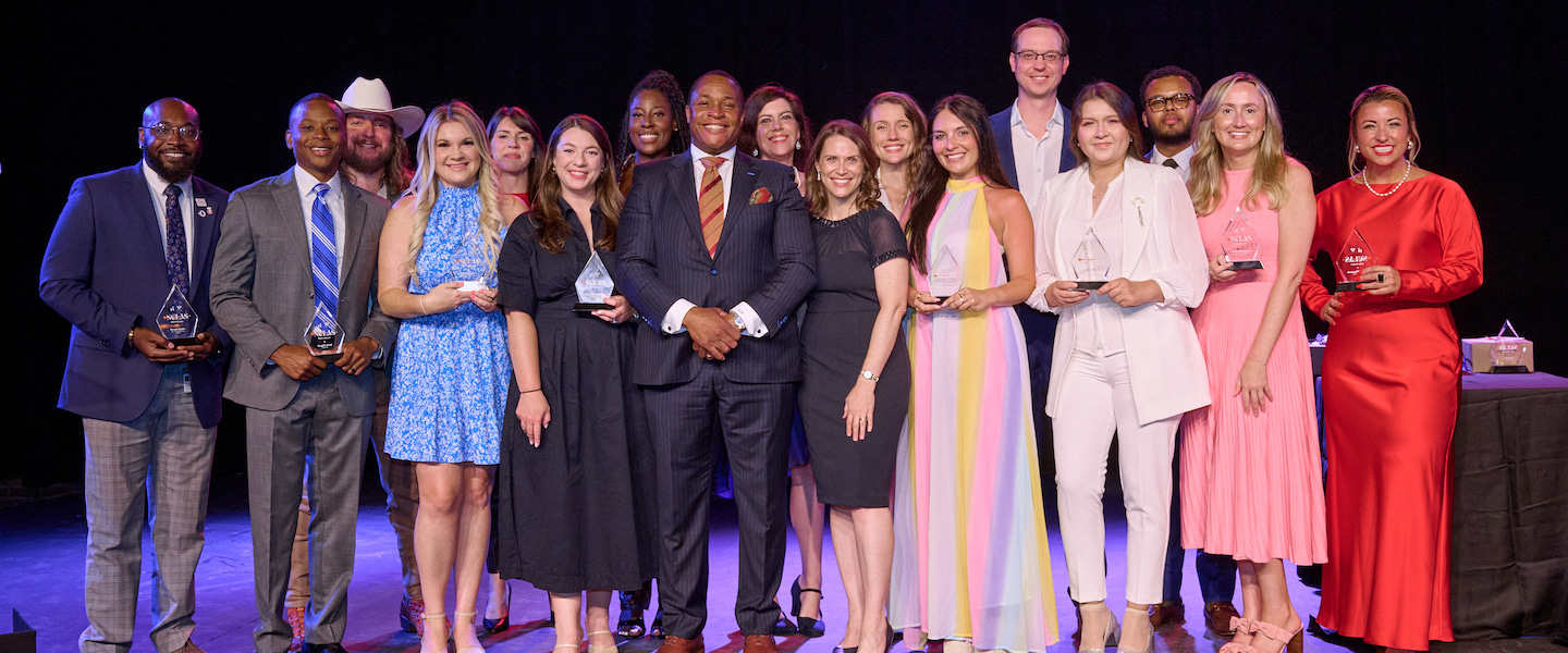 Group of award winners pose on stage, smiling and holding trophies in formal attire