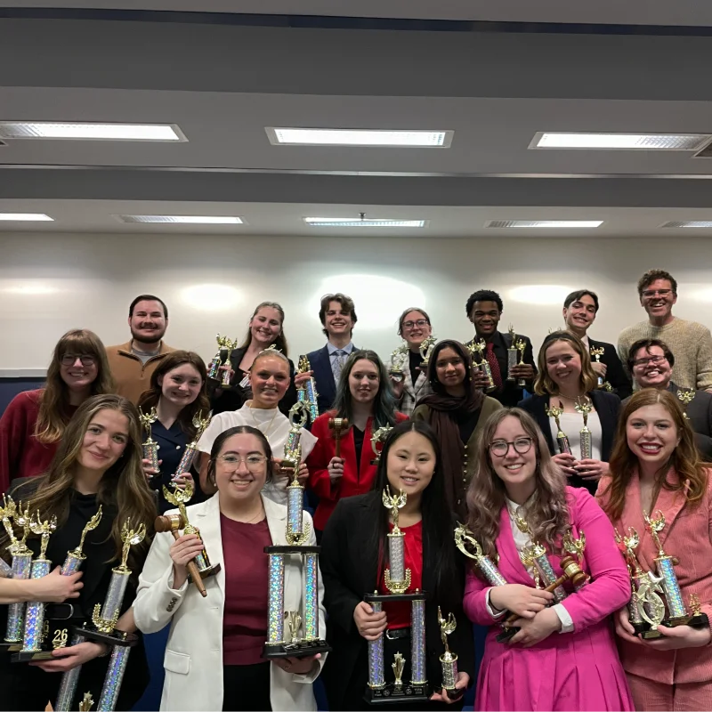 Group of smiling students proudly holding many trophies after winning an academic competition.