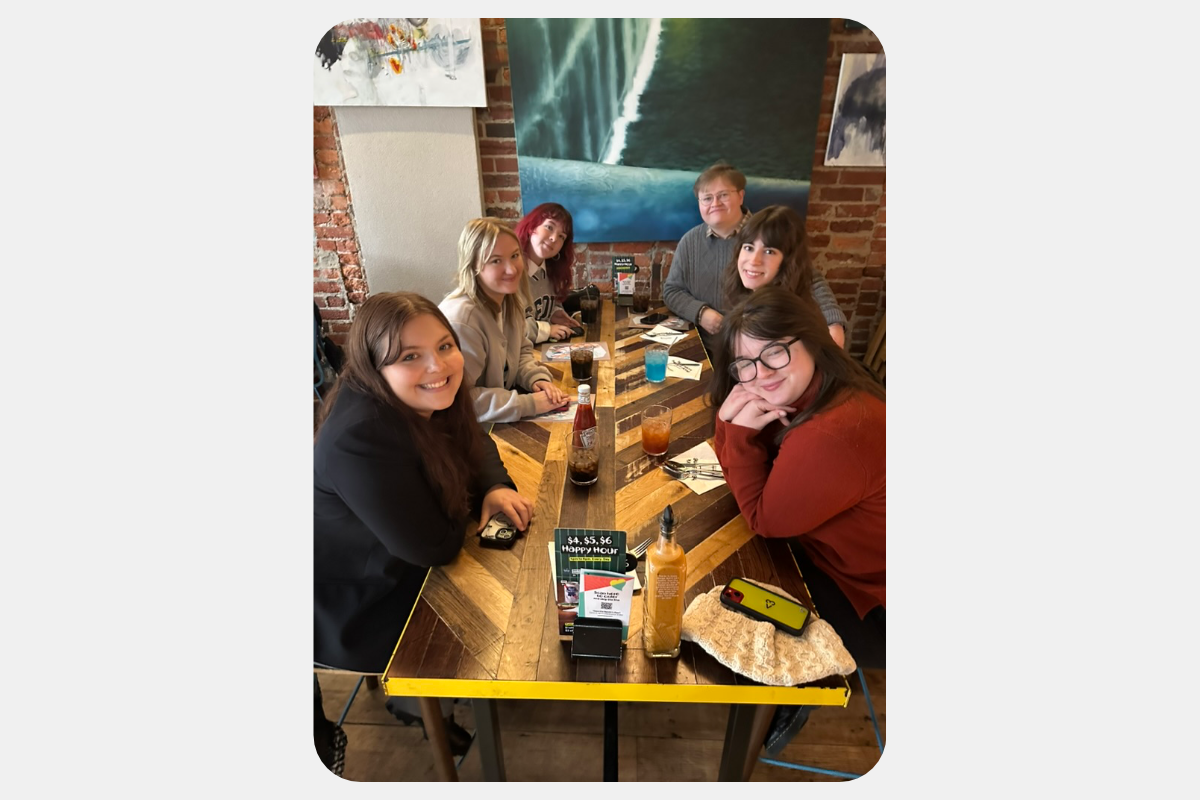 Six happy friends enjoying a meal at a rustic restaurant table, smiling for the camera.