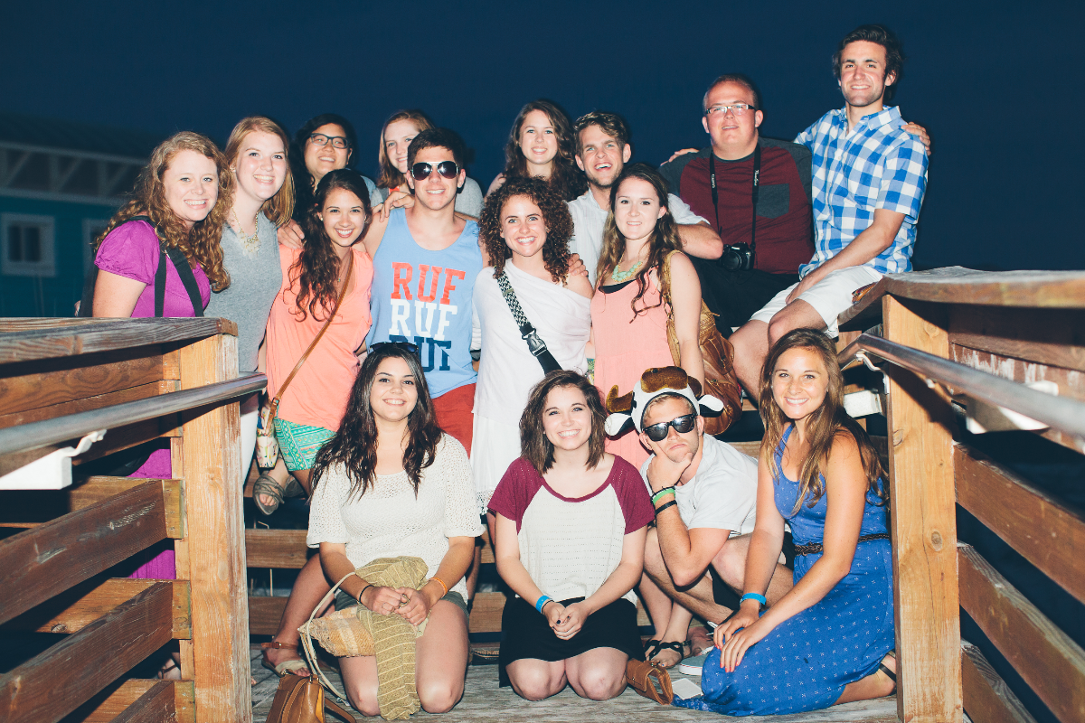 Large group of smiling young adults and friends posing on a wooden deck at night, enjoying a social gathering.