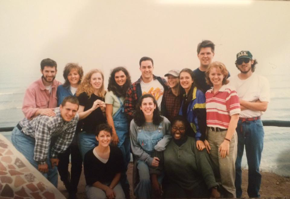 Diverse group of young adults smiling outdoors by the ocean, friends on a coastal trip.