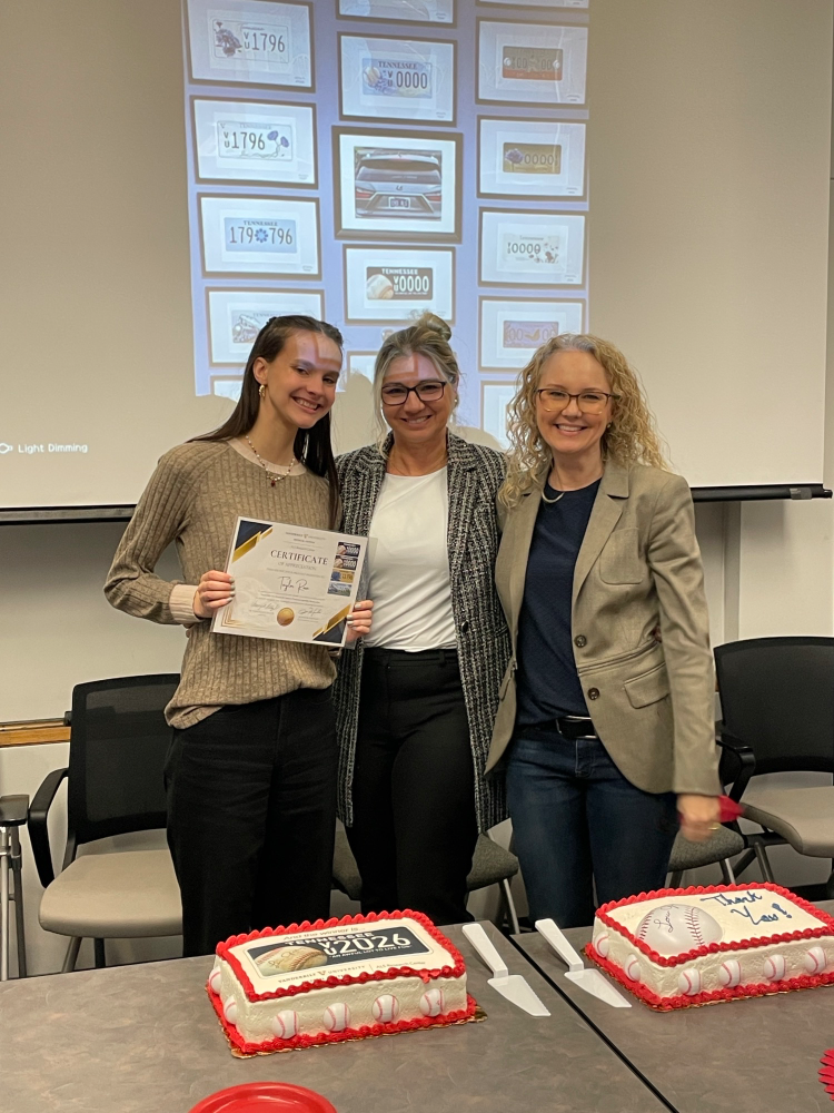 Young woman receives award certificate, flanked by two women, with baseball-themed celebration cakes.