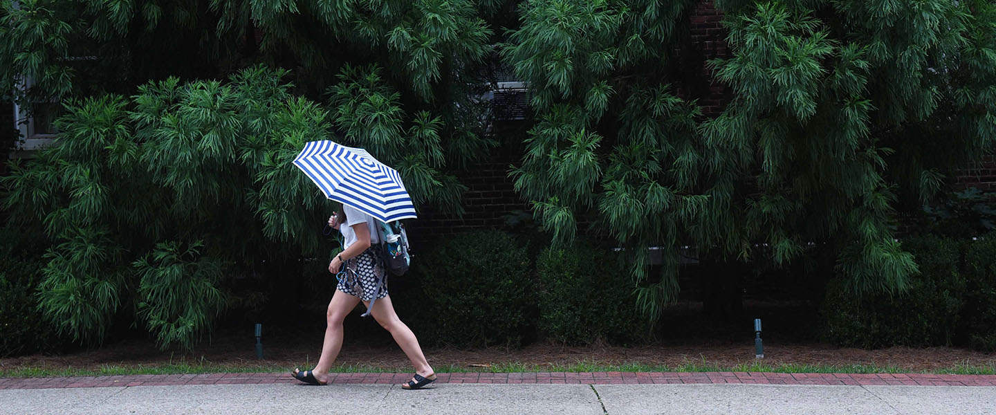 A student walking in the rain with an umbrella with green foliage in the background