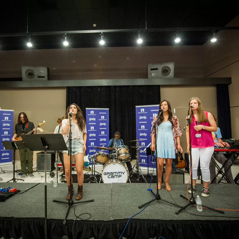 Young musicians perform on a stage at Grammy Camp. Three vocalists sing into microphones, backed by a drummer, guitarist, and keyboardist, creating a lively and energetic atmosphere.