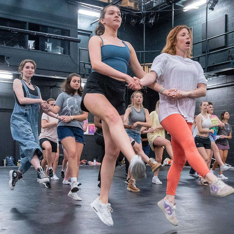 A group of students participate in a high-energy dance rehearsal in a black box theater. They are holding hands and performing synchronized high kicks, led by an instructor wearing bright orange leggings and a white shirt. The students, dressed in casual athletic and dancewear, display focus and determination as they follow the choreography. The background features black walls, stage lighting, and various props, indicating a dynamic performing arts environment.