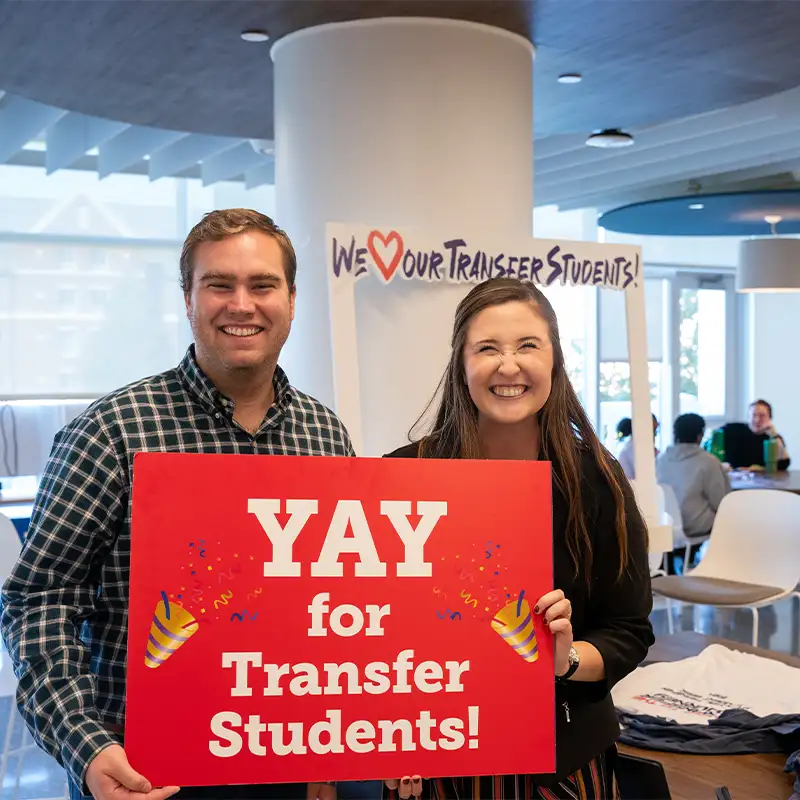 Two smiling individuals, a man and a woman, stand together in a bright space holding a large red sign that reads "YAY for Transfer Students!" The background features a decorative sign saying "We ❤️ Our Transfer Students!" and people sitting at tables.